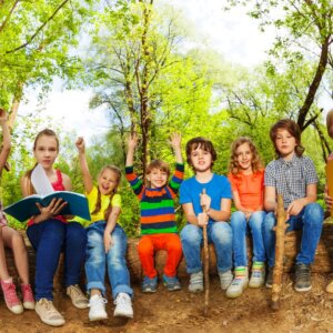 Bambini sorridenti seduti su tronchi nel bosco, con libri e quaderni colorati, circondati dalla natura. Un momento di gioia e condivisione che riflette lo spirito del Summer Camp Sancy 2025 – dove divertimento, sport e amicizia crescono insieme.